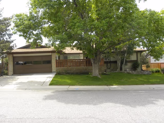 Front exterior view of a single-story residential building with a brown garage door, a wooden porch railing, and a large tree with green leaves in the front yard. The lawn is well-maintained and there is a sidewalk and street in front of the house.