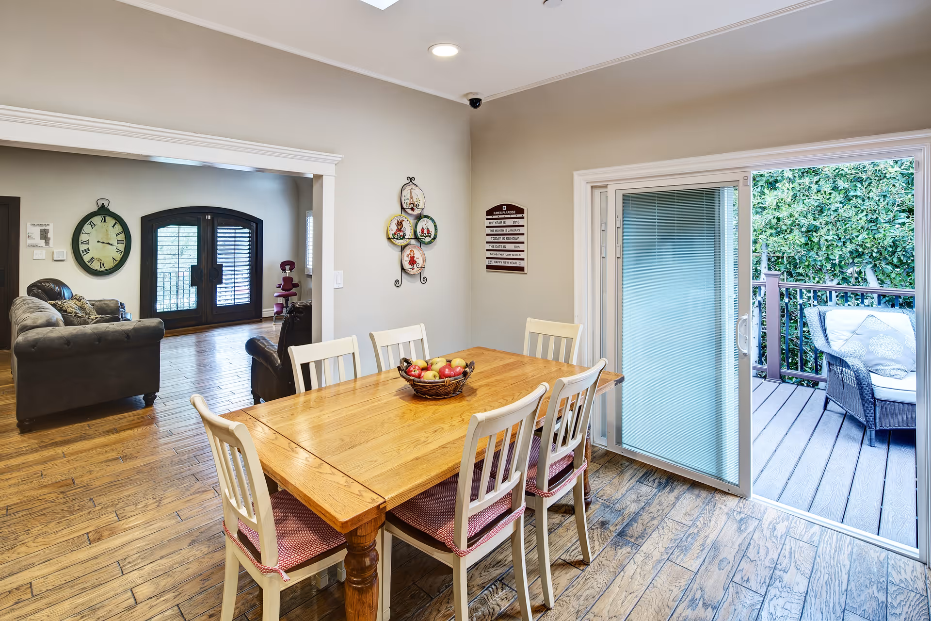 A dining area with a wooden table and six white chairs with red cushions. A basket of apples is placed on the table. To the right, there is a sliding glass door leading to an outdoor patio with a cushioned wicker chair and greenery. In the background, a living room area is visible with a gray sofa, a large wall clock, and double doors with wooden shutters.