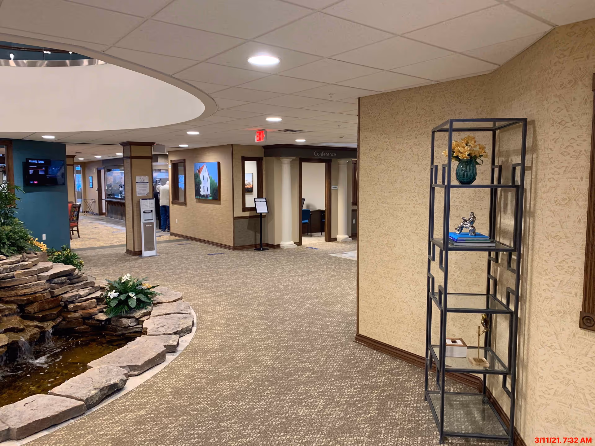 Interior view of a hallway in a senior living facility with a stone water feature on the left, a metal shelf with decorative items on the right, and a conference room entrance straight ahead. The area is well-lit with ceiling lights and has carpeted flooring.