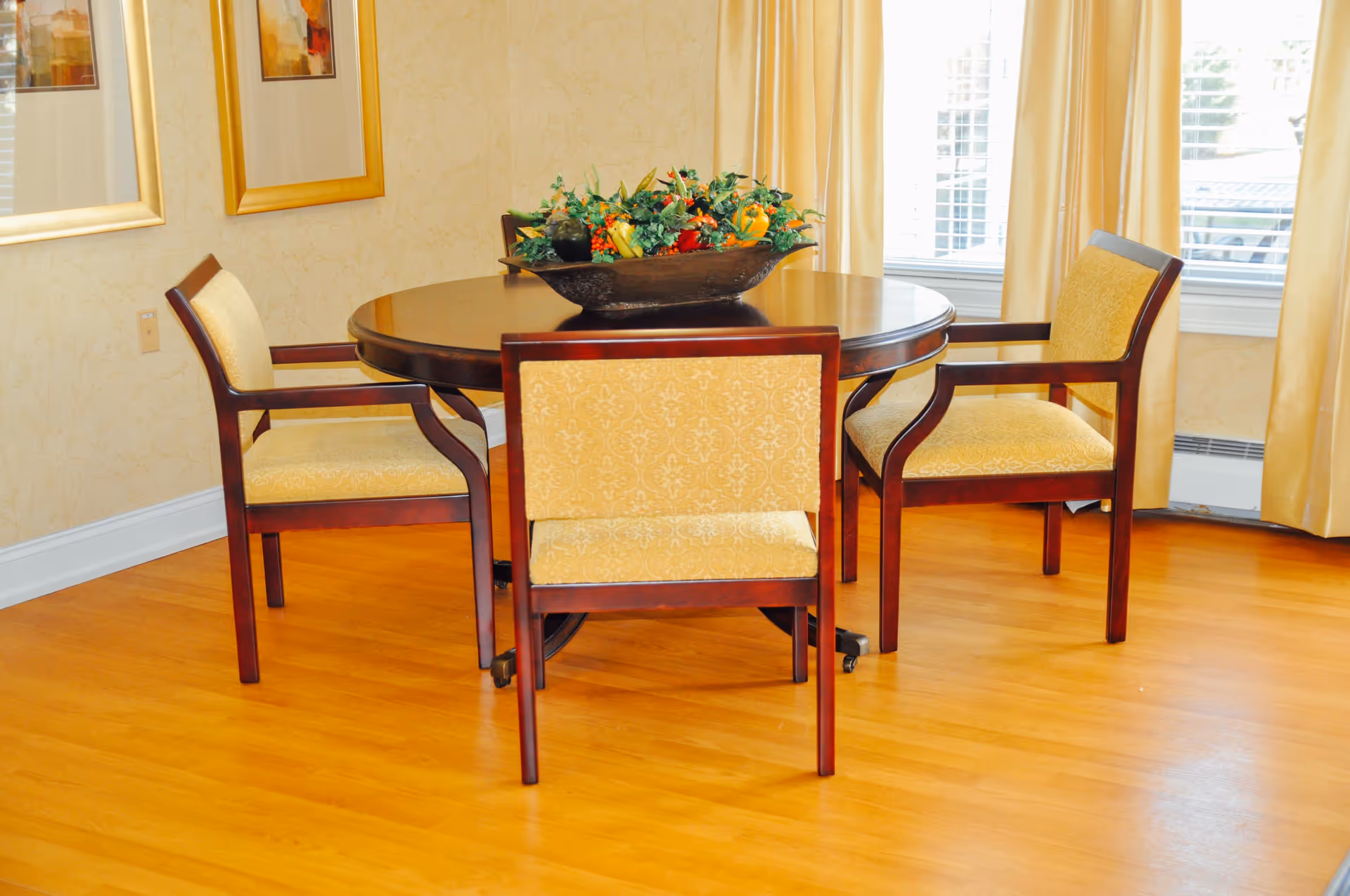 Round wooden dining table with four upholstered chairs and a floral centerpiece in a well-lit room with hardwood floors and curtains.