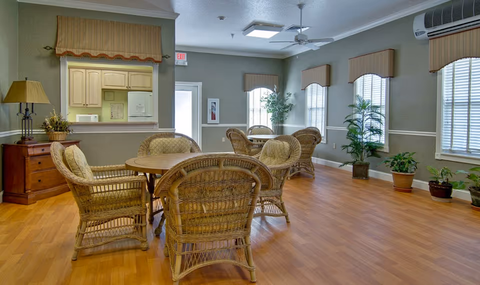 Sunlit common room with round wooden tables surrounded by wicker chairs, hardwood floor, potted plants, and windows with valances.