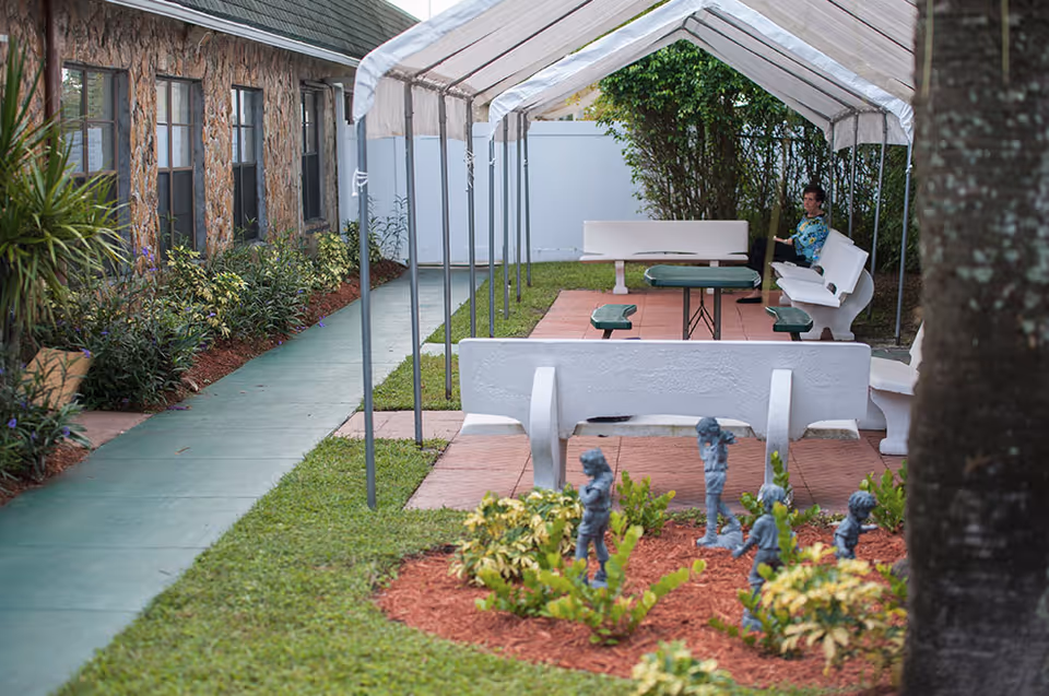 Outdoor courtyard with covered picnic tables and white benches, a small garden with statues, and a person sitting on a bench.