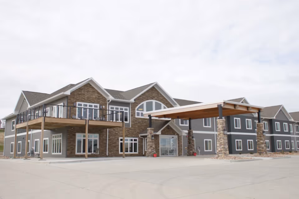 Exterior view of a two-story senior living facility building with stone and gray siding, large windows, a covered entrance supported by stone pillars, and a balcony on the second floor.