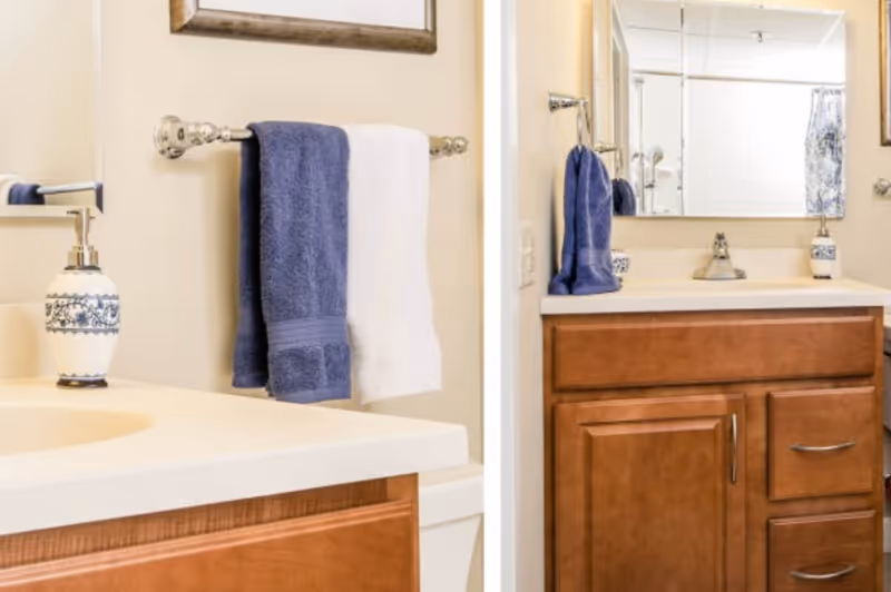A bathroom vanity with a wooden cabinet, a white countertop, a sink, and a large mirror above. There are two towels hanging on a silver towel rack, one blue and one white. A decorative soap dispenser with a blue and white pattern is placed on the countertop.