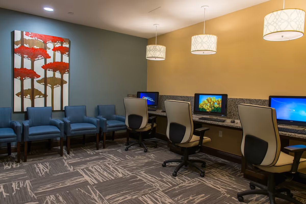 A computer room with three desktop computers on a granite countertop, each with an office chair. To the left, there are five blue upholstered chairs arranged against a green wall with a large painting of red and brown trees. The room has patterned carpet and three hanging pendant lights with white lampshades.