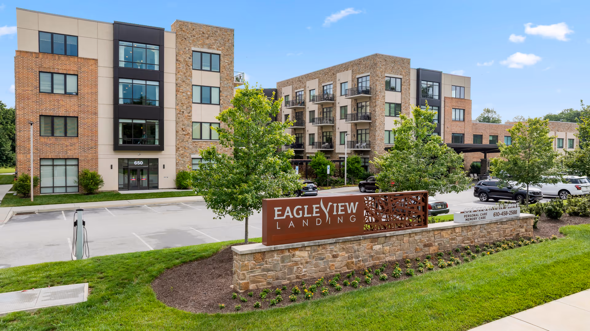 Exterior view of Eagleview Landing senior living facility showing a modern multi-story building with brick and stone facade, balconies, parking lot, green lawn, trees, and a large sign with the facility name in front.