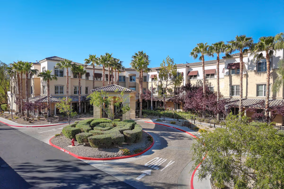 Exterior view of a multi-story senior living facility with beige walls, red-tiled roofs, and numerous palm trees surrounding the building. There is a circular driveway with a landscaped roundabout featuring trimmed bushes and a fire hydrant. The sky is clear and blue.