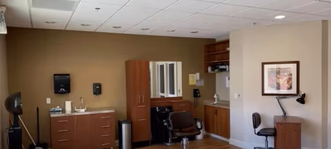 Interior view of a room with wooden cabinets, a sink, a mirror, and chairs. The room has beige walls, a framed picture on the wall, and a ceiling with recessed lighting.