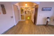 View of a hallway entrance in a senior living facility with double doors open leading to a well-lit corridor. The floor is wooden, and there is a pineapple wall decoration on the left and a sign that reads 'FRANZEN' on the right wall above a small decorative item.