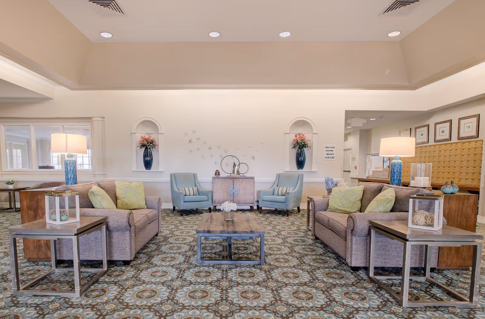 A spacious and well-lit living room area in a senior living facility featuring two beige sofas with green pillows, two blue armchairs, a wooden coffee table, and side tables with lamps. The room has patterned carpet flooring, decorative wall niches with vases of flowers, and wall art of birds in flight. Mailboxes and framed pictures are visible on the right side of the room.