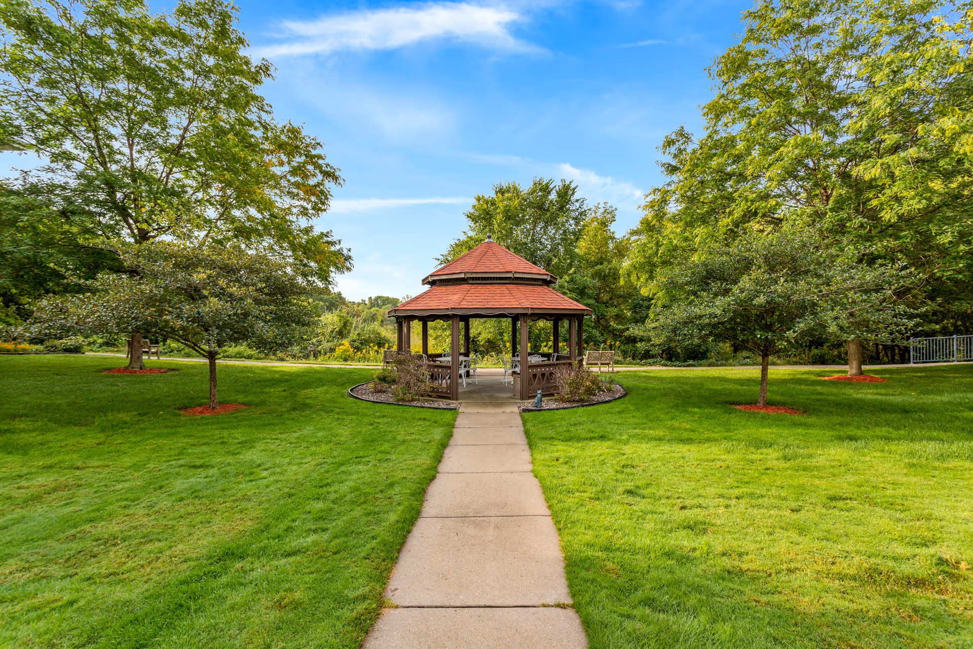 A paved walkway leading to a wooden gazebo with a red roof, surrounded by green grass and trees under a blue sky with some clouds.