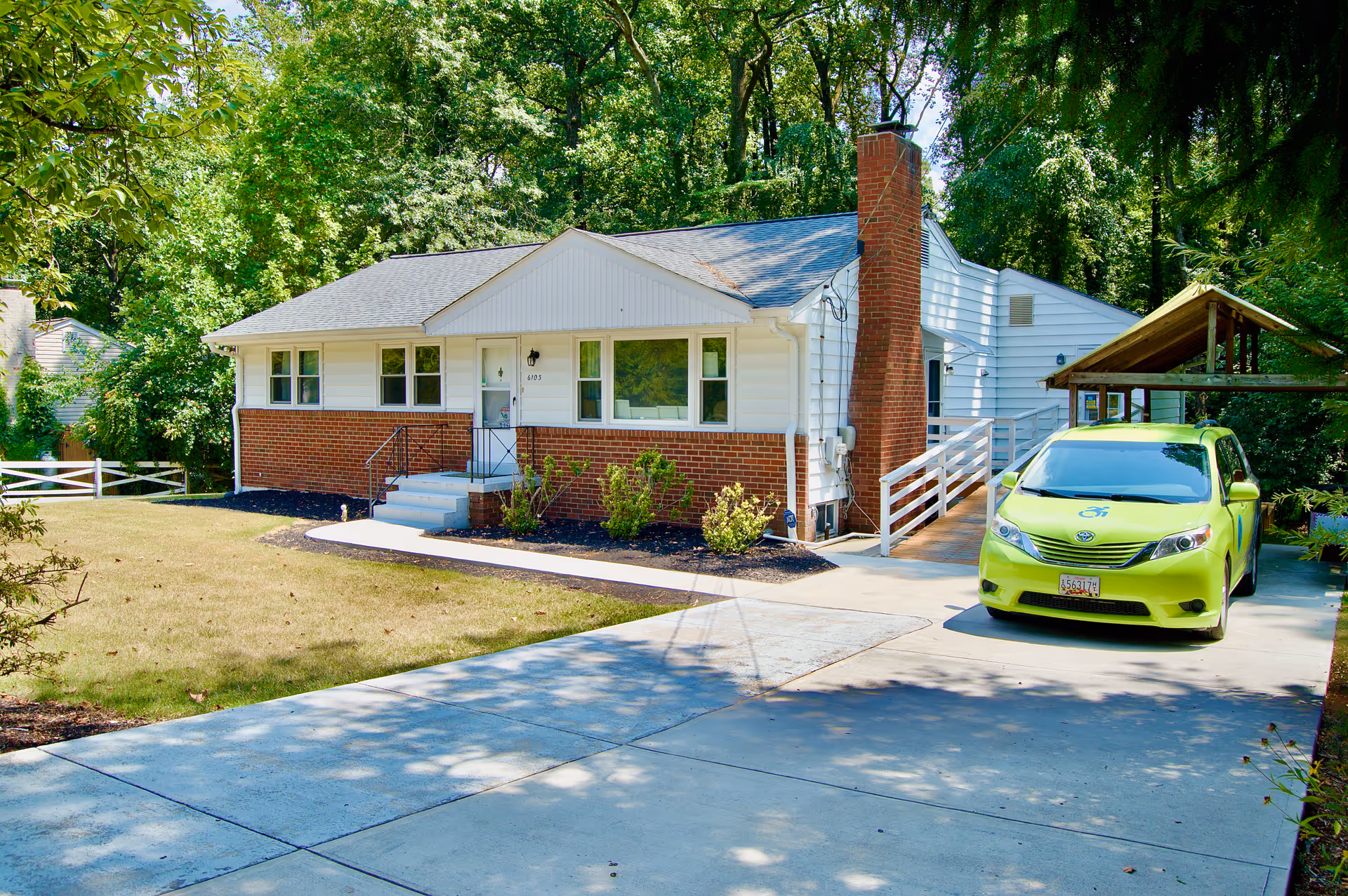 A single-story house with white siding and red brick exterior, surrounded by trees and greenery. There is a concrete driveway leading to the house, with a bright green car parked under a wooden carport on the right side. The house has a small front porch with steps and a ramp for accessibility.