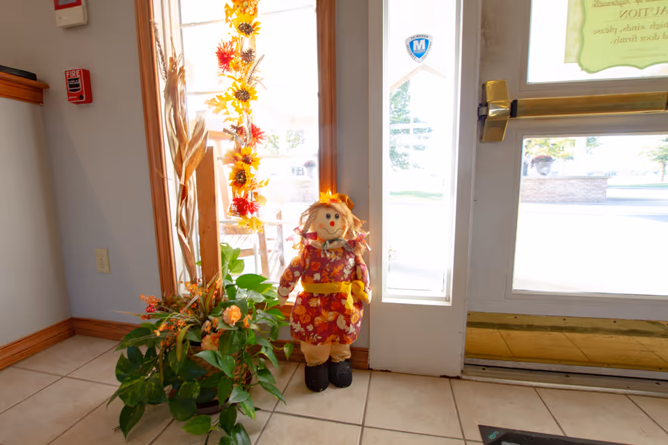 Indoor corner near an entrance door with a decorative scarecrow doll dressed in fall-themed clothes, a green potted plant with orange flowers, and a vertical garland of autumn leaves hanging on the window. A fire alarm and electrical outlet are visible on the wall.
