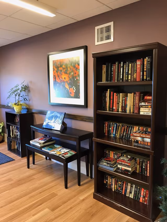 Interior of a room with wooden flooring and purple walls featuring a tall dark wooden bookshelf filled with books, a smaller bookshelf with books and plants on top, and two nested tables with magazines and a framed picture of a snowy mountain scene. A framed floral artwork hangs on the wall above the tables.