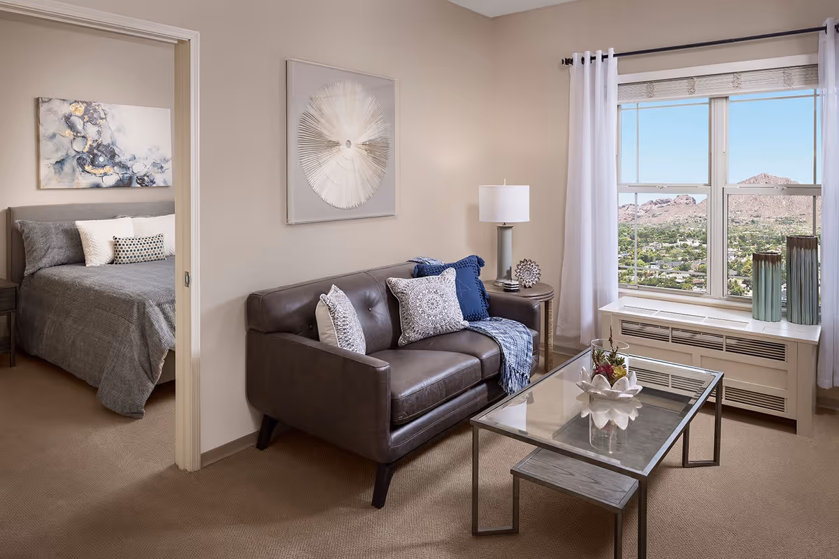 A cozy living room with a dark brown leather loveseat adorned with decorative pillows and a throw blanket. A glass coffee table with a floral centerpiece sits in front of the loveseat. To the right, a large window with white curtains offers a scenic view of a mountainous landscape. A side table with a lamp and decorative items is next to the loveseat. In the background, an open doorway reveals a bedroom with a bed covered in gray bedding and pillows, and a piece of abstract art hanging above the bed.