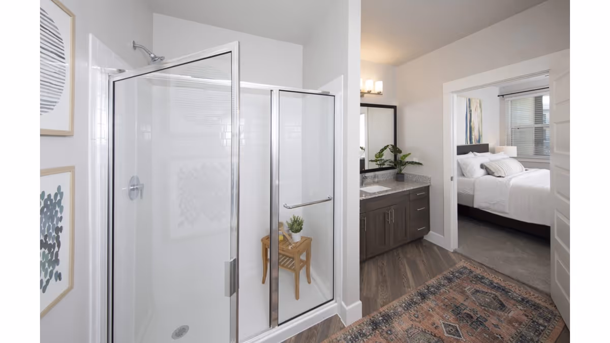 A modern bathroom with a glass-enclosed shower featuring a small wooden stool with a plant on it. Adjacent to the shower is a vanity with a granite countertop, a sink, a large mirror, and a potted plant. The bathroom opens into a bedroom with a neatly made bed, white bedding, and a window with blinds. The floor has wood-style flooring with a patterned rug near the entrance to the bedroom.