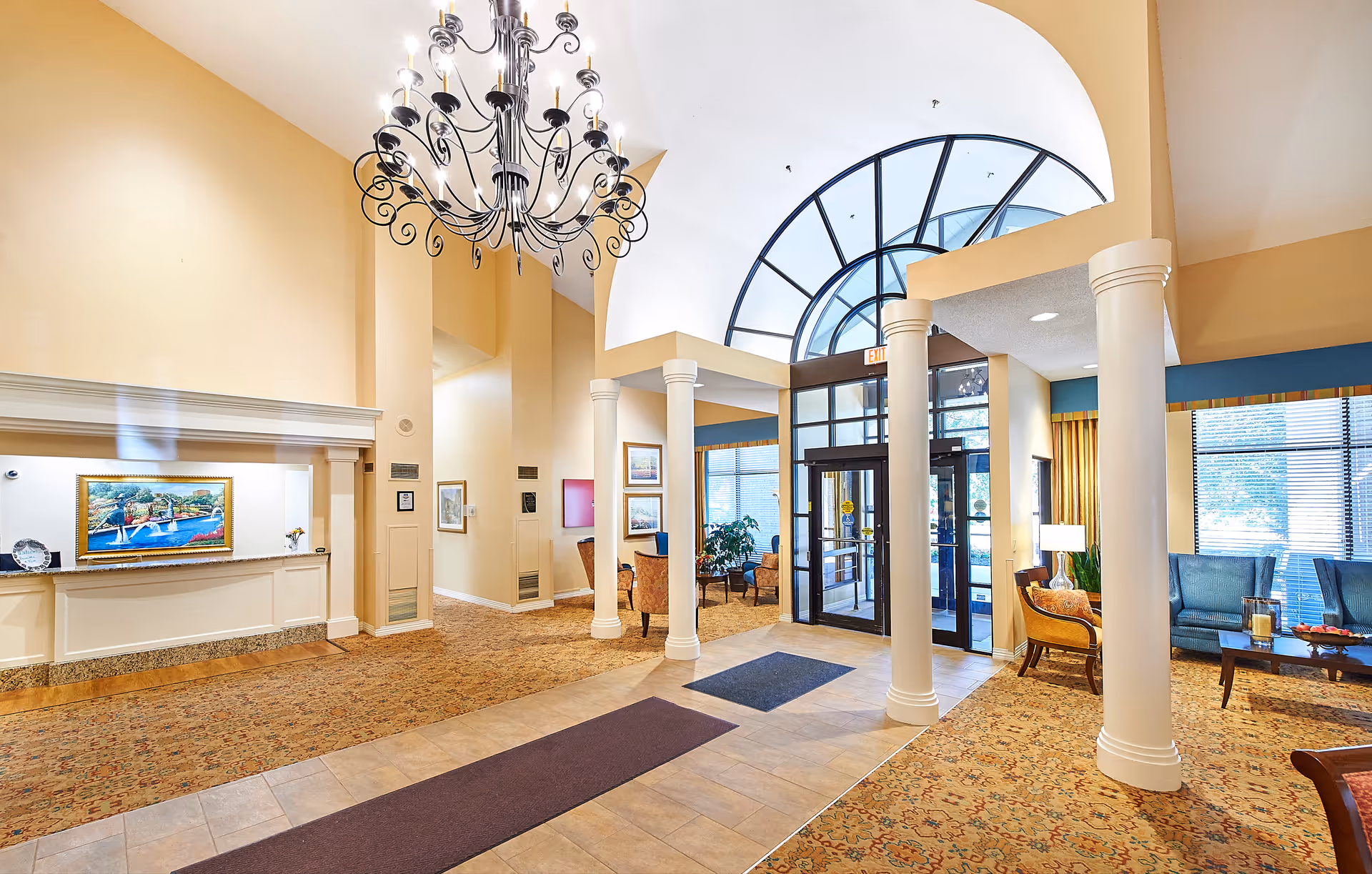 Bright and spacious senior living facility lobby with a large arched window above the entrance doors, white columns, a decorative chandelier, seating areas with chairs and tables, and a reception desk with a colorful painting behind it.