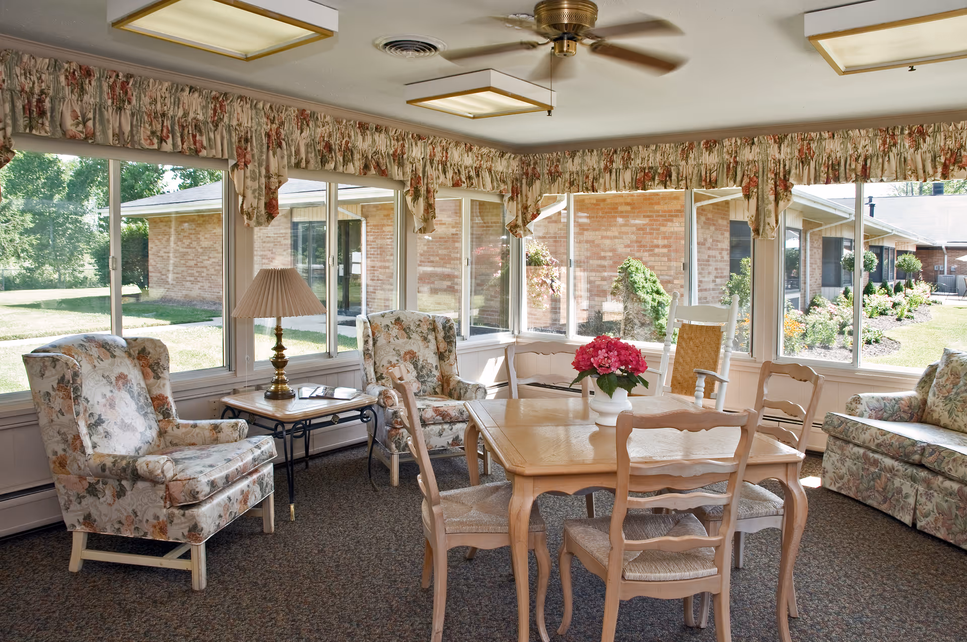 Sunlit common sitting room with floral upholstered chairs, a wooden table topped with a vase of pink flowers, and large windows overlooking a courtyard.