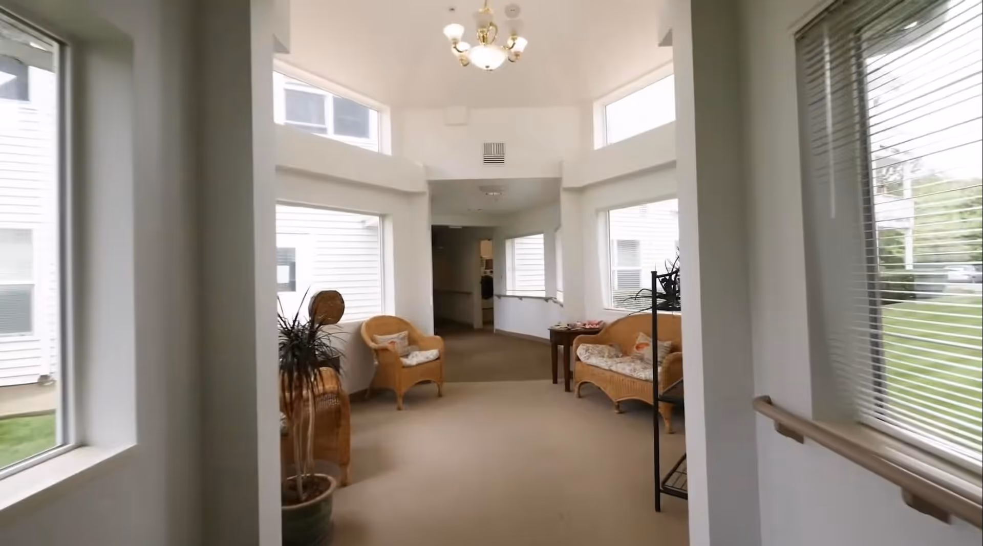 Bright interior common area with wicker chairs, side tables, large windows, and a handrail along a carpeted corridor.