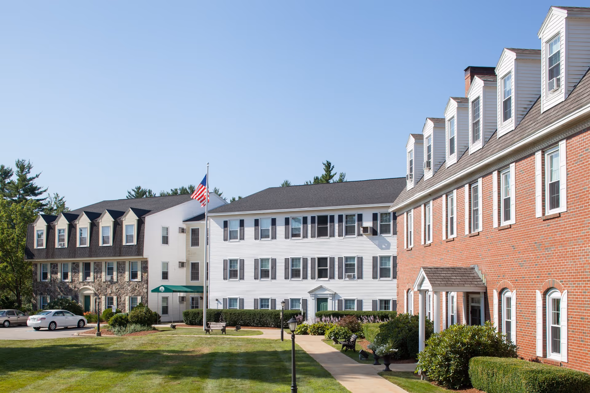 Exterior view of Langdon Place of Exeter, showing a multi-story building with a combination of brick, stone, and white siding. There is a flagpole with an American flag in the center, a well-maintained lawn, benches, shrubs, and a few parked cars under a clear blue sky.