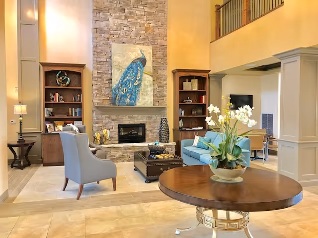 Spacious, well-lit living room with a stone fireplace topped by a peacock painting, seating area, bookshelves and a round table with flowers.
