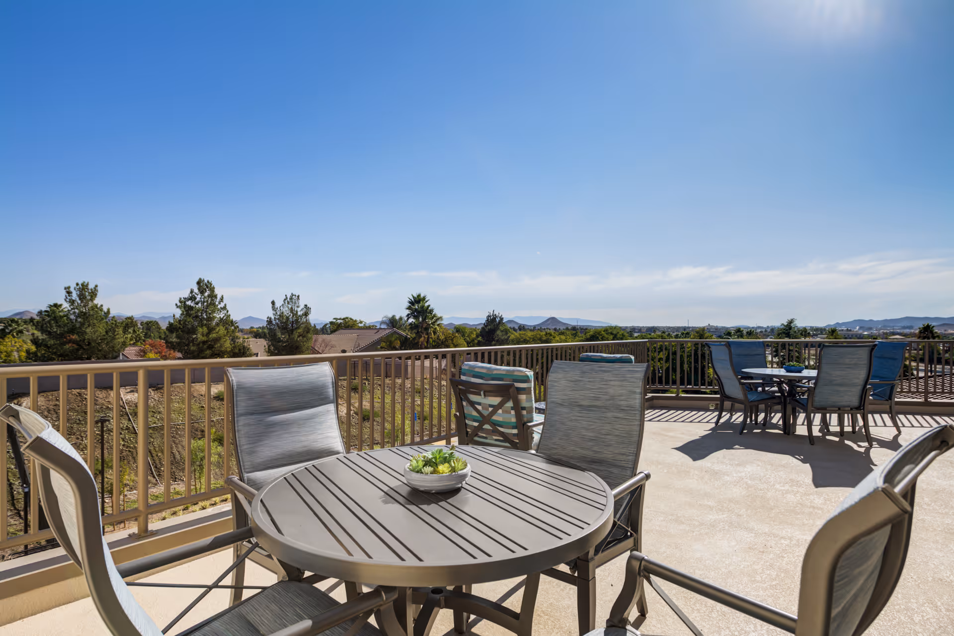 Outdoor patio area with round tables and chairs under a clear blue sky, overlooking trees, rooftops, and distant mountains.