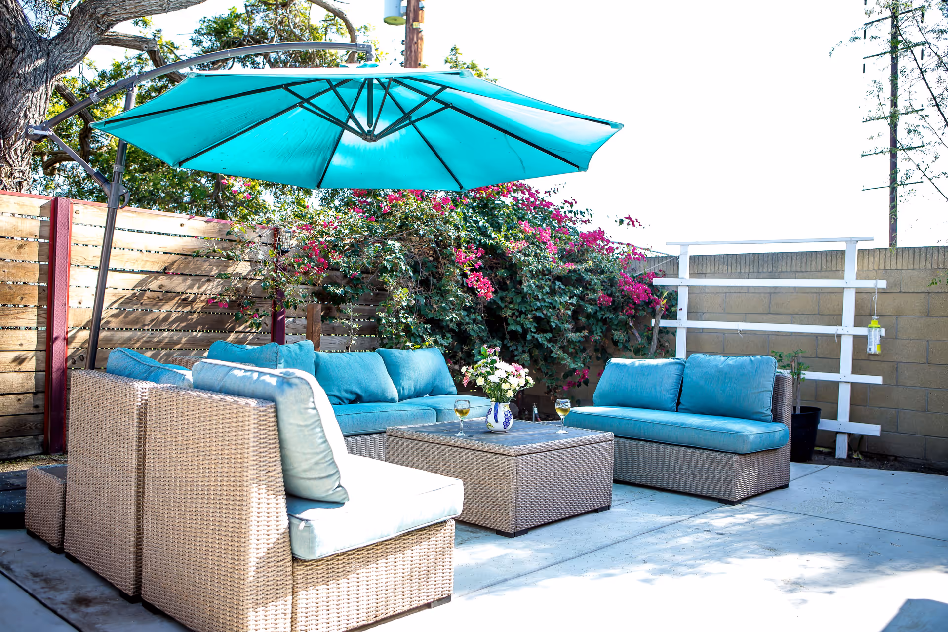 Outdoor patio area with wicker furniture including a sofa, loveseat, and chair with blue cushions. A large turquoise umbrella provides shade. There is a coffee table with a vase of flowers and two glasses of white wine. The area is surrounded by a wooden fence and a wall with flowering plants and greenery.