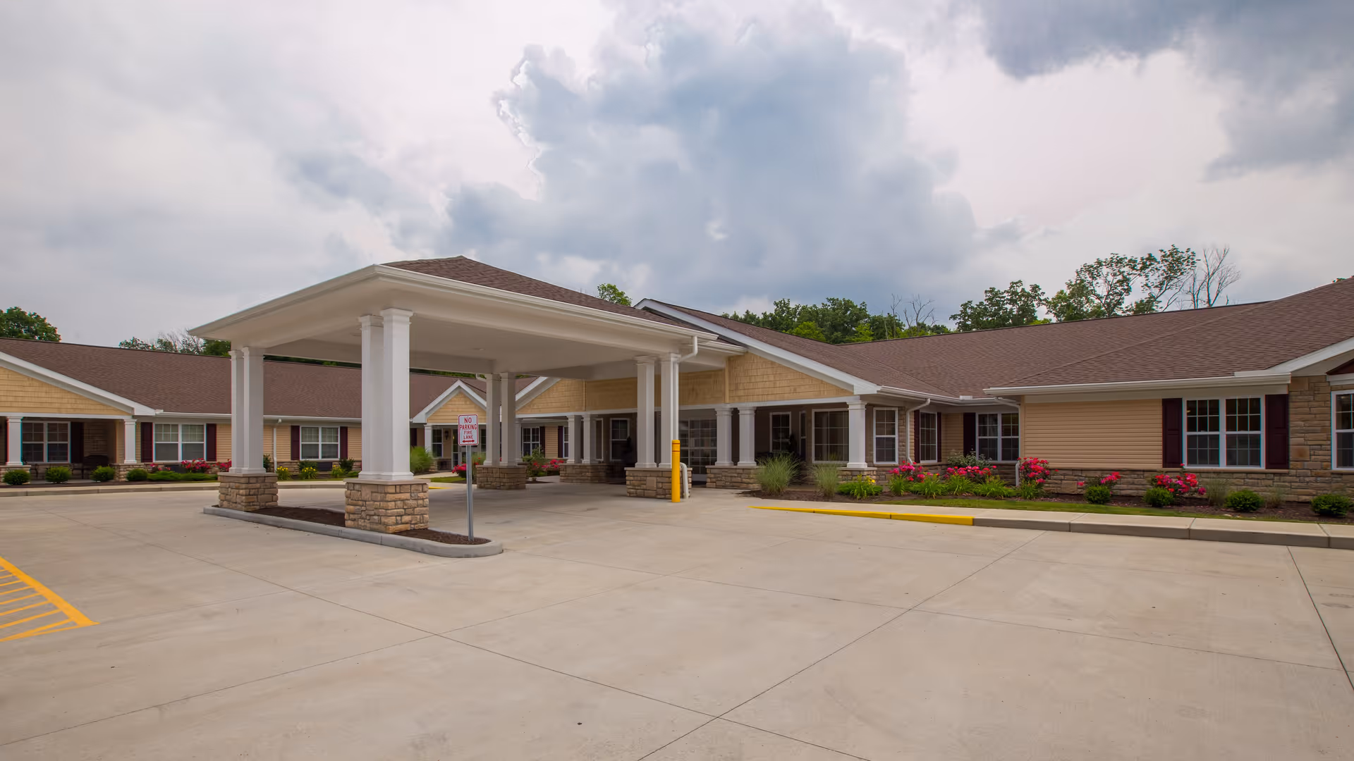 Exterior view of a single-story senior living facility building with a covered entrance supported by white columns and stone bases. The building has beige siding, brown roof shingles, and several windows with burgundy shutters. There are landscaped flower beds with pink flowers and green shrubs along the building. The sky is cloudy.
