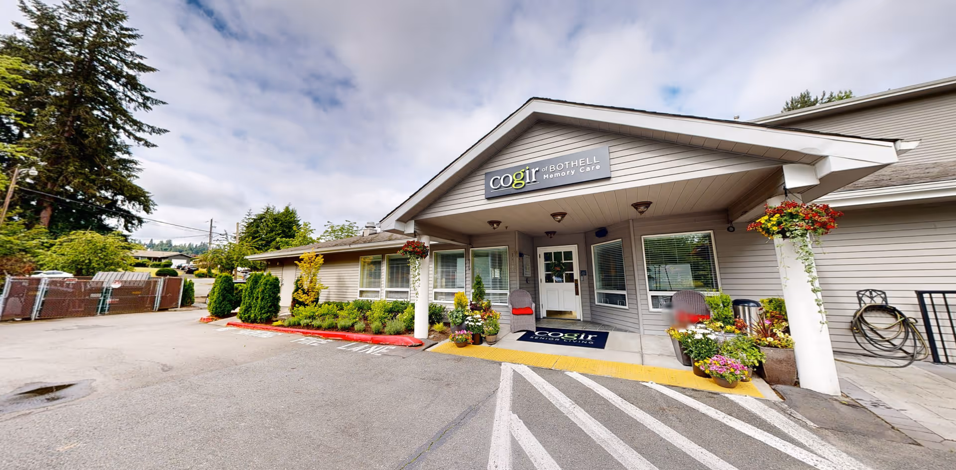 Exterior view of Cogir of Bothell Memory Care facility entrance with a covered porch, potted plants, and a welcome mat. The building is light gray with white trim, and there are bushes and trees around the parking area under a partly cloudy sky.