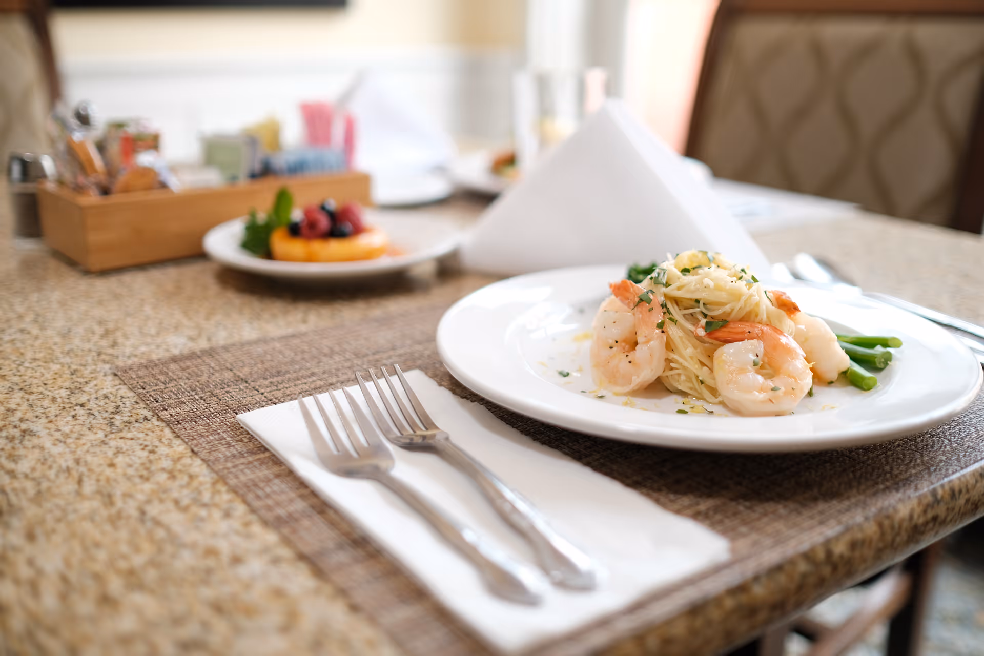 A close-up view of a dining table set with a plate of shrimp pasta garnished with herbs and green beans. In the background, there is a plate with a fruit tart and a wooden container holding condiments and utensils. Two forks rest on a white napkin beside the main plate.