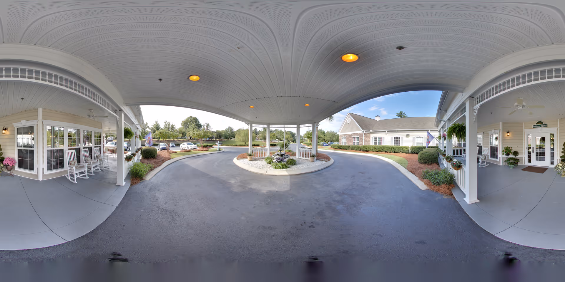 Covered driveway entrance of The Legacy of Lexington senior living facility with white rocking chairs on the left porch, potted plants, and a circular flower bed in the center. The building exterior is light-colored with large windows and a welcoming entrance on the right side. Several cars are parked in the background under a partly cloudy sky.