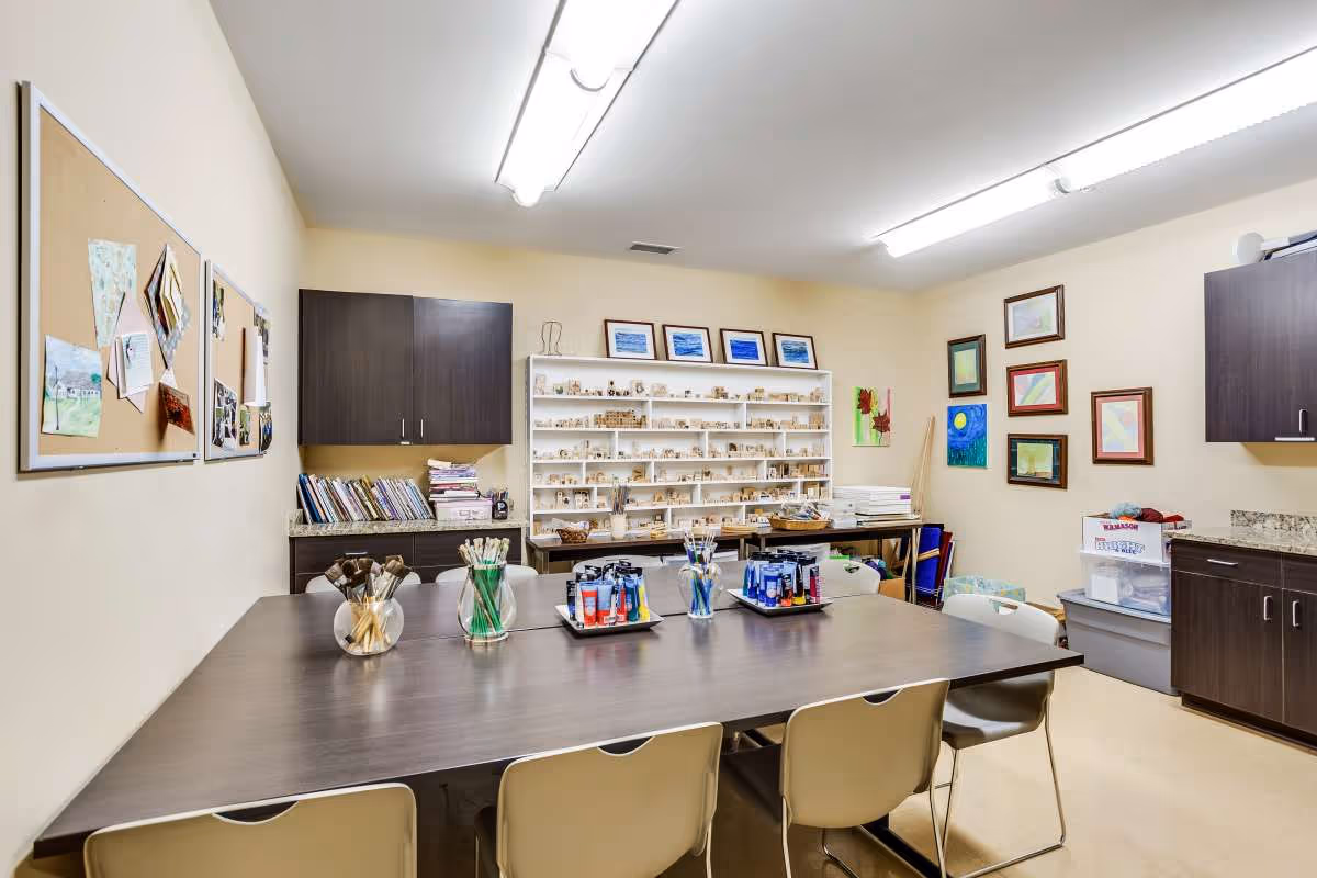 A well-lit craft room with a large dark wooden table surrounded by beige chairs. On the table are jars filled with paintbrushes and trays holding various paint tubes. The walls are decorated with framed artwork and bulletin boards with pinned papers. Shelves and cabinets hold books, craft supplies, and small wooden blocks.