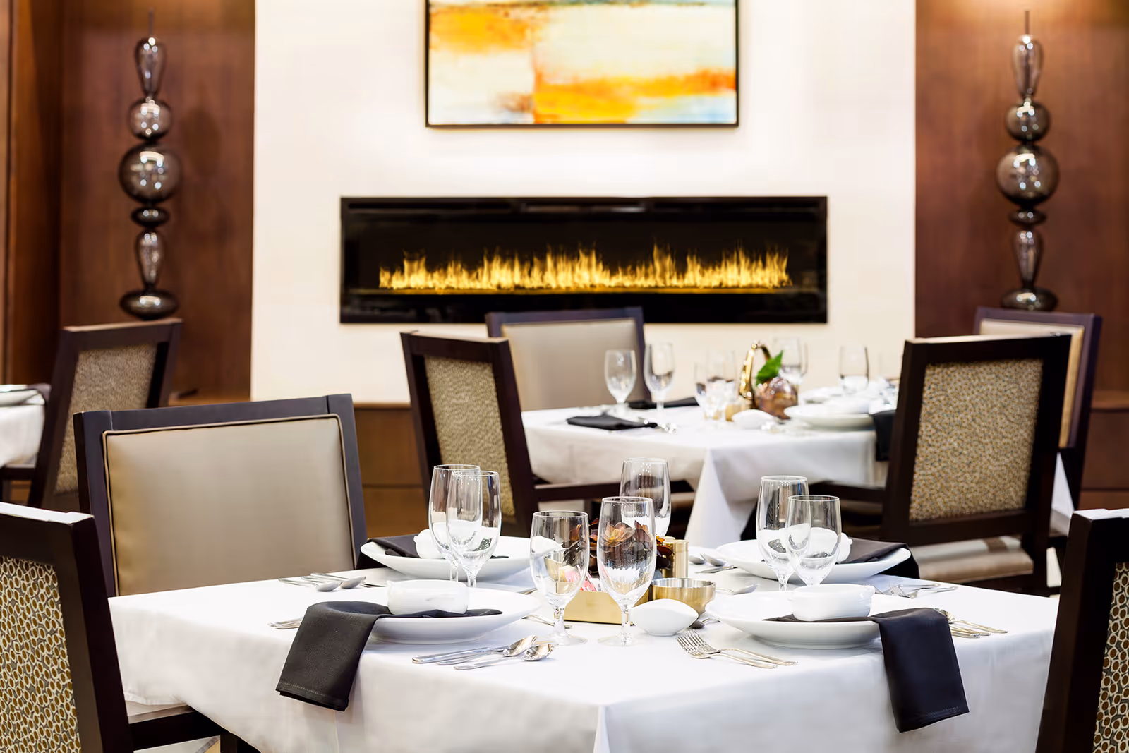 A dining room with tables set for a meal, featuring white tablecloths, neatly arranged plates, glasses, silverware, and black napkins. In the background, there is a modern rectangular fireplace with flames and a colorful abstract painting above it. The chairs have dark wooden frames with beige and patterned upholstery.