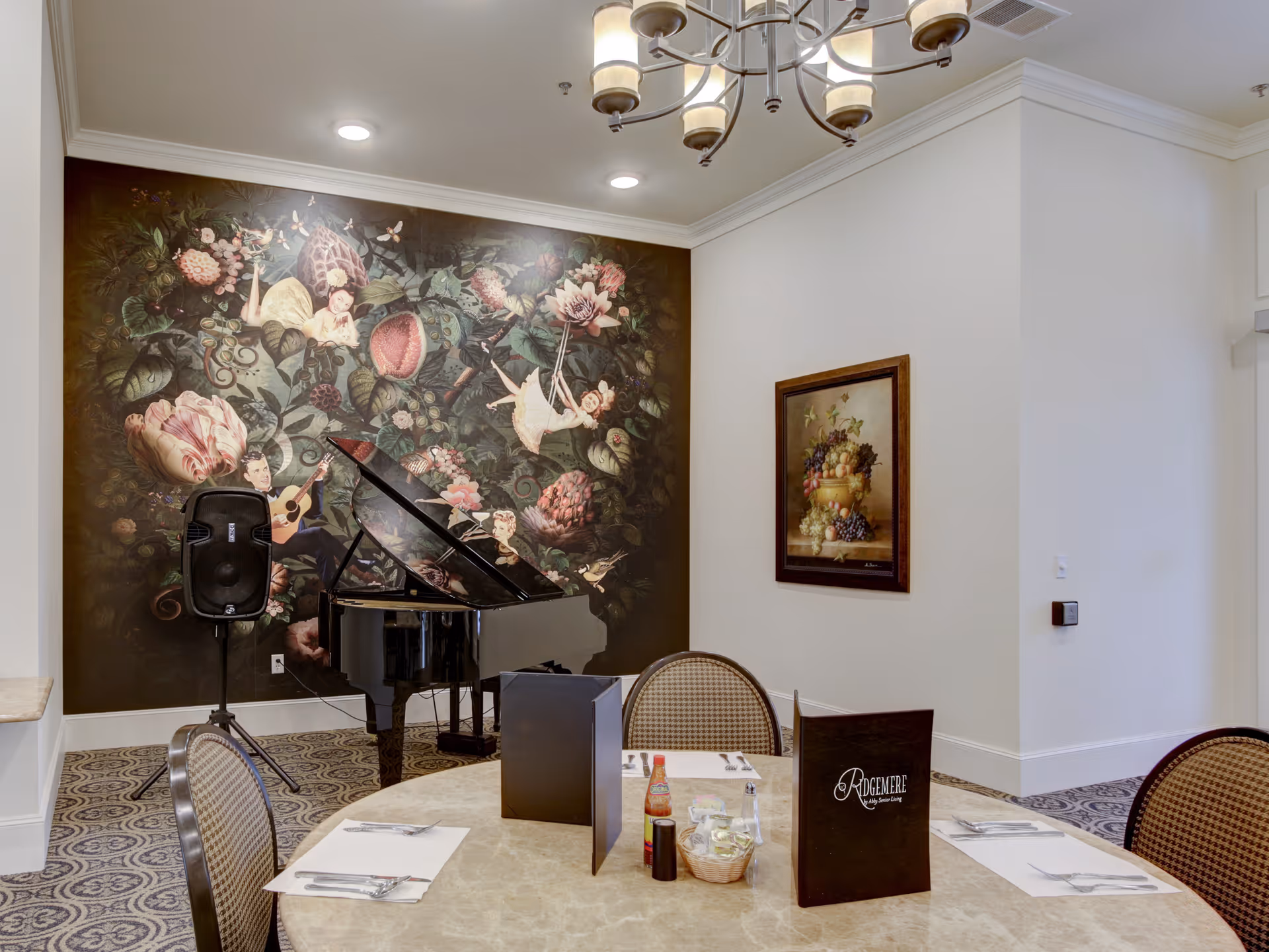 Dining room with a round table set with menus and place settings, a grand piano in front of a large floral mural, and a chandelier overhead.