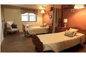 A senior living facility bedroom with two single beds, each with beige bedding and pillows. There is a window with curtains, a white armchair with a pillow, a bedside table with a lamp, and warm-colored walls with framed pictures.