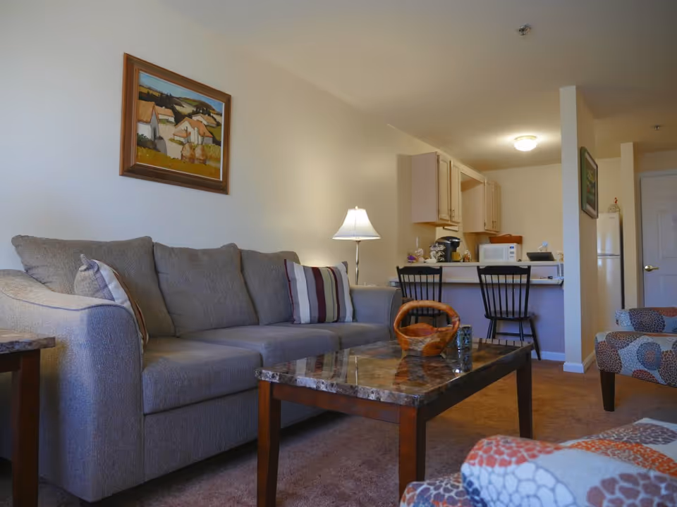 A cozy living room area with a gray sofa adorned with striped pillows, a marble-top coffee table with a decorative basket, and a patterned armchair. In the background, there is a small kitchen with wooden cabinets, a microwave, a coffee maker, and two black bar stools at the counter. A framed painting hangs on the wall above the sofa, and a floor lamp provides soft lighting.