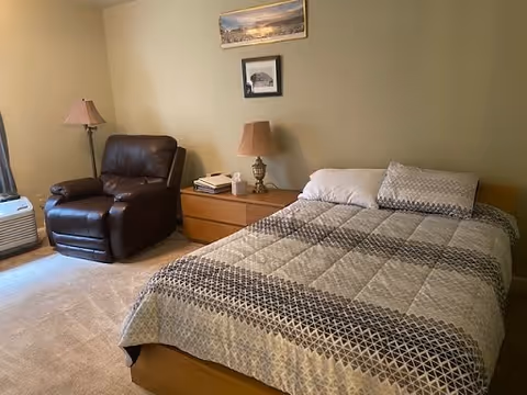 A cozy bedroom with a double bed covered in a patterned quilt, a wooden nightstand with a lamp and some books, and a dark brown leather armchair next to a window with curtains. Two framed pictures hang on the beige wall above the nightstand.