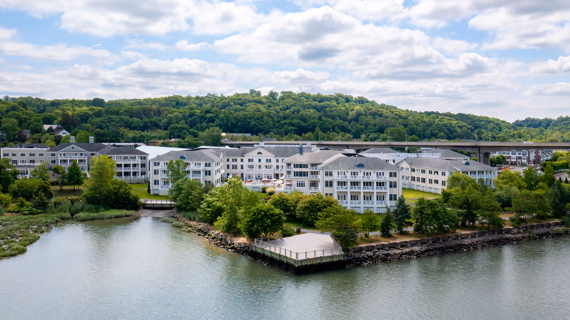 Atria on Roslyn Harbor senior living facility situated along a waterfront with a dock, surrounded by trees and greenery, with a bridge and forested hills in the background under a partly cloudy sky.