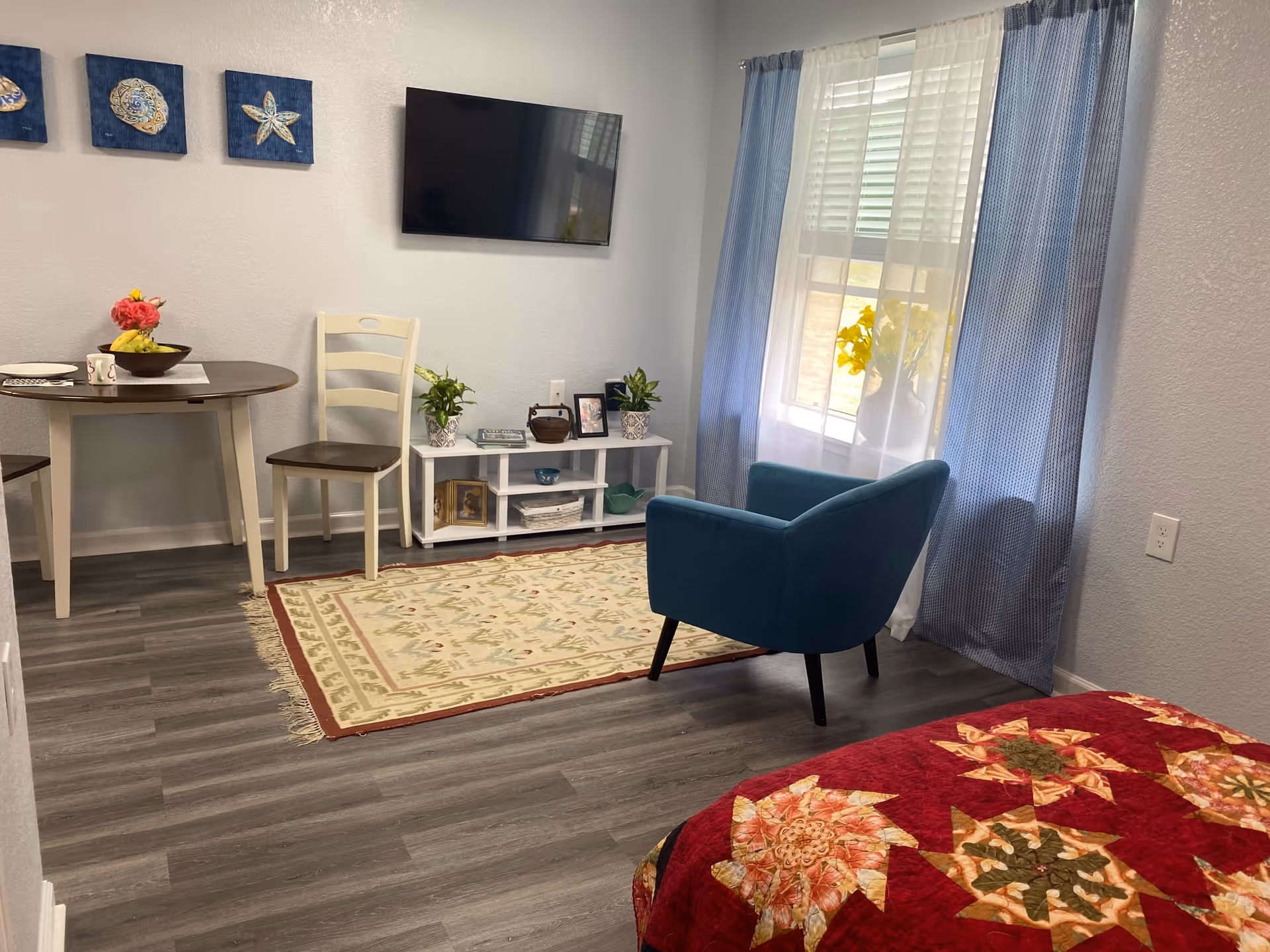 A cozy room with a small dining table and two chairs on the left, a blue armchair near a window with blue curtains on the right, a white TV stand with plants and picture frames underneath a wall-mounted flat screen TV, and a bed with a red floral quilt partially visible in the foreground.