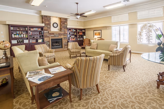 A bright and spacious living room with beige patterned carpet, multiple upholstered chairs and sofas arranged around a wooden coffee table with magazines. The room features built-in wooden bookshelves flanking a stone fireplace with a clock above it. Large windows with white blinds allow natural light to fill the space, and a ceiling fan is mounted above.
