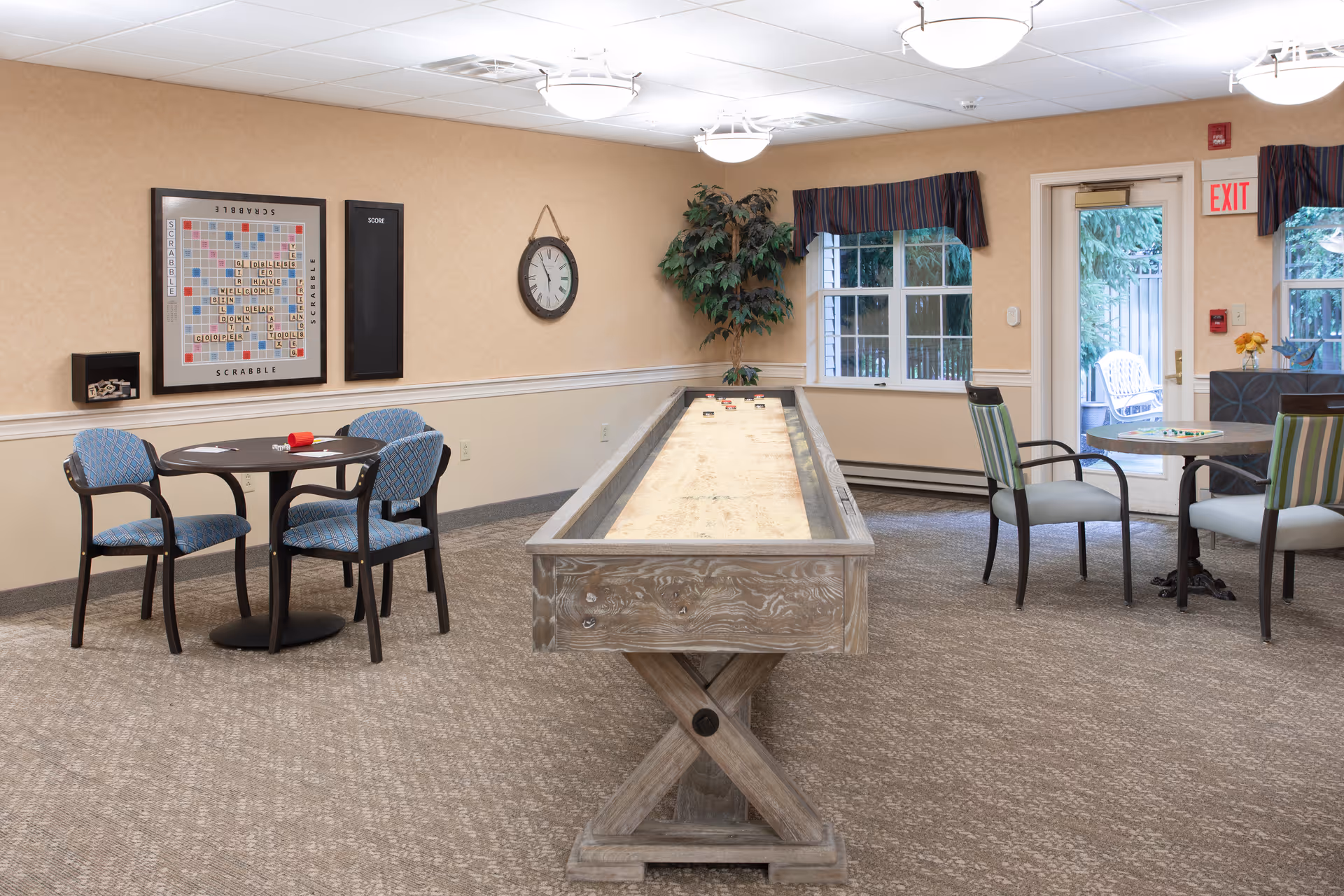 A well-lit game room in Governor's Village featuring a shuffleboard table in the center, two round tables with chairs on either side, a large Scrabble board mounted on the wall, a clock, and a potted plant near windows and a glass door leading outside.