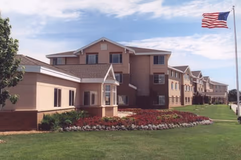 Exterior view of a multi-story senior living facility building with beige walls and brown roof accents, surrounded by a well-maintained lawn and flower beds. An American flag flies on a tall flagpole to the right under a partly cloudy sky.