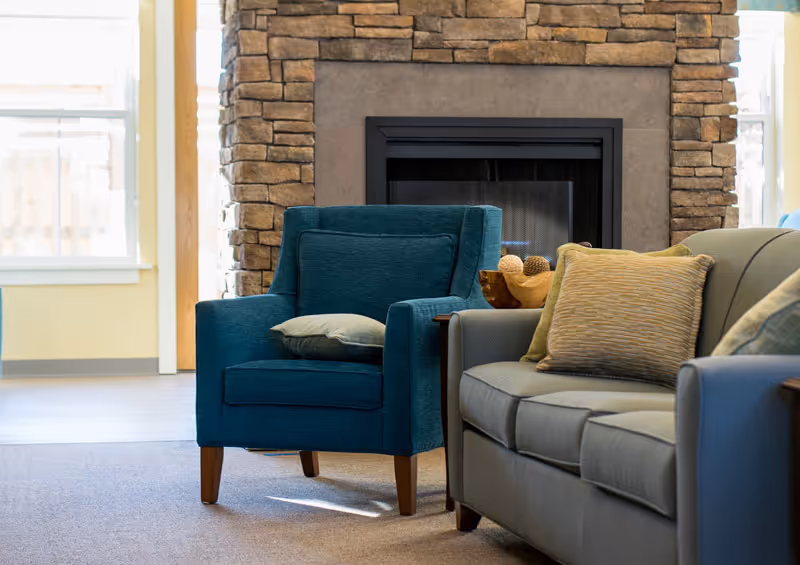 A cozy living room area featuring a teal armchair with a cushion, a gray sofa with multiple pillows, and a stone fireplace in the background. Natural light comes through windows on either side of the fireplace.