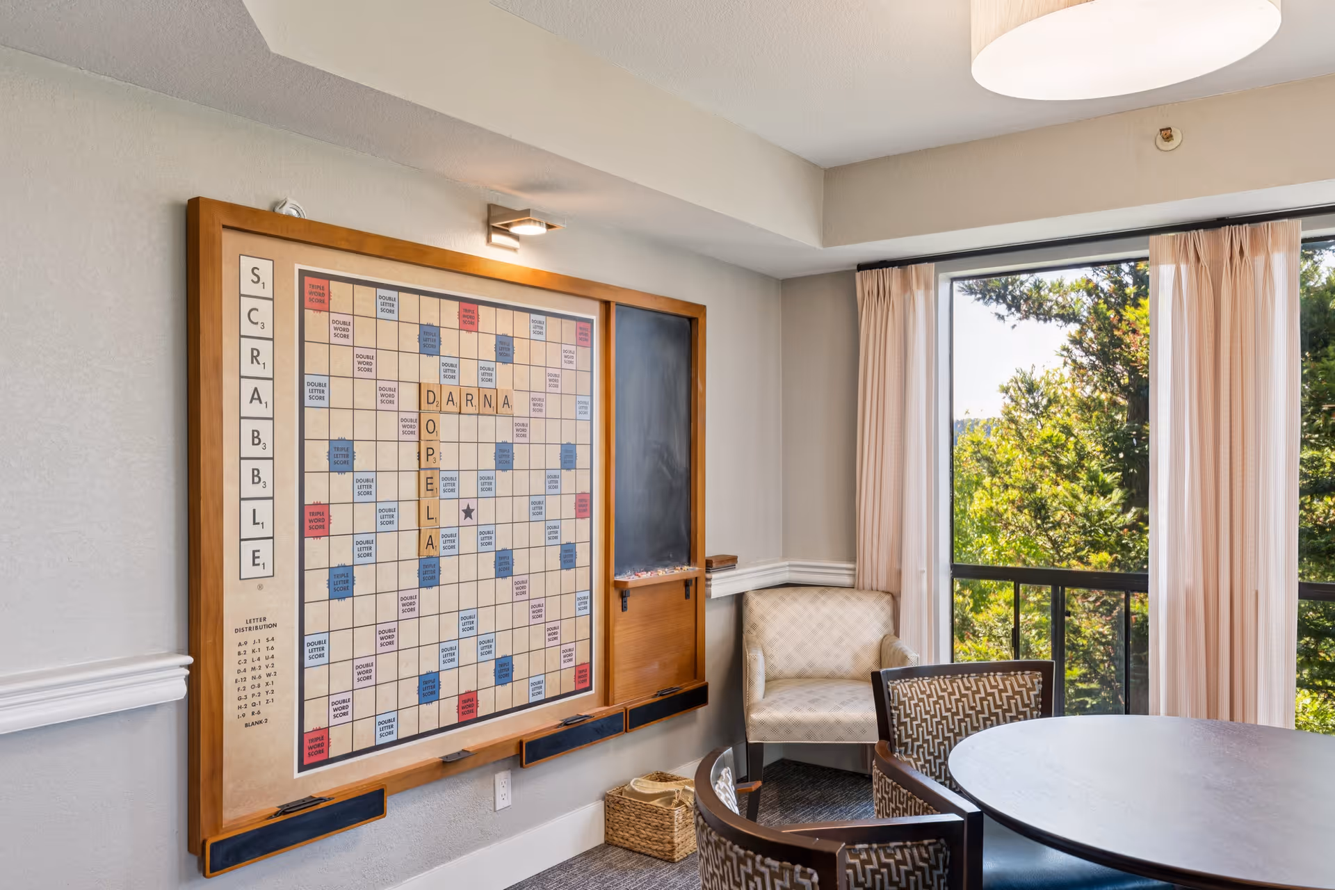 A cozy room with a large wall-mounted Scrabble board, a small chalkboard, a round table with patterned chairs, an armchair, and a large window with curtains showing green trees outside.