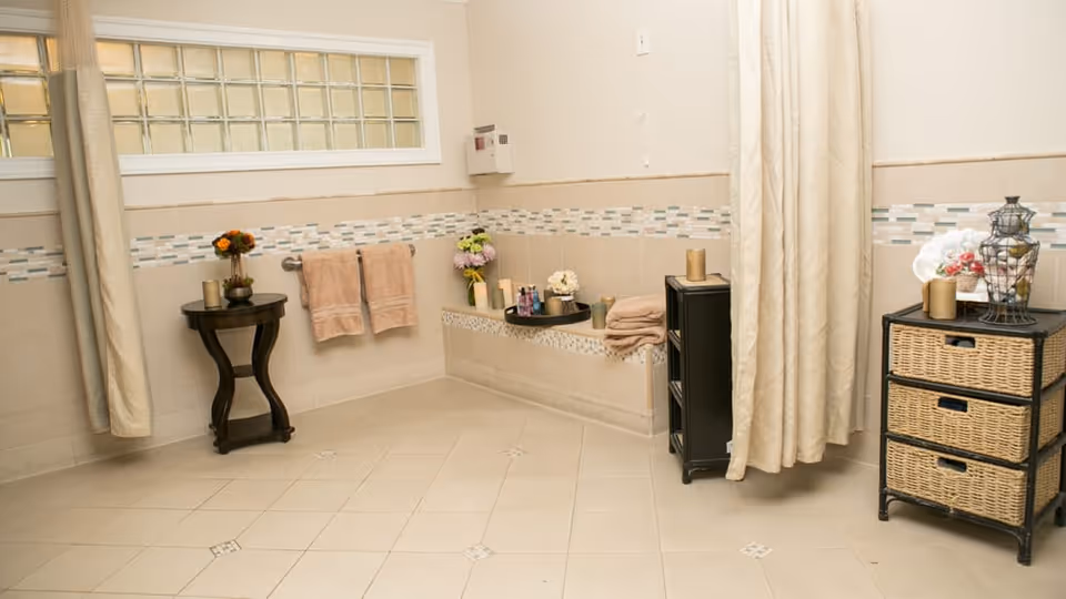 A clean and spacious bathroom with beige tiled floors and walls, featuring a small black side table with a flower vase, two beige towels hanging on a towel rack, a built-in bench with toiletries and folded towels, a black storage unit with more towels, and a wicker drawer unit with decorative items. A beige shower curtain is partially visible on the right side.