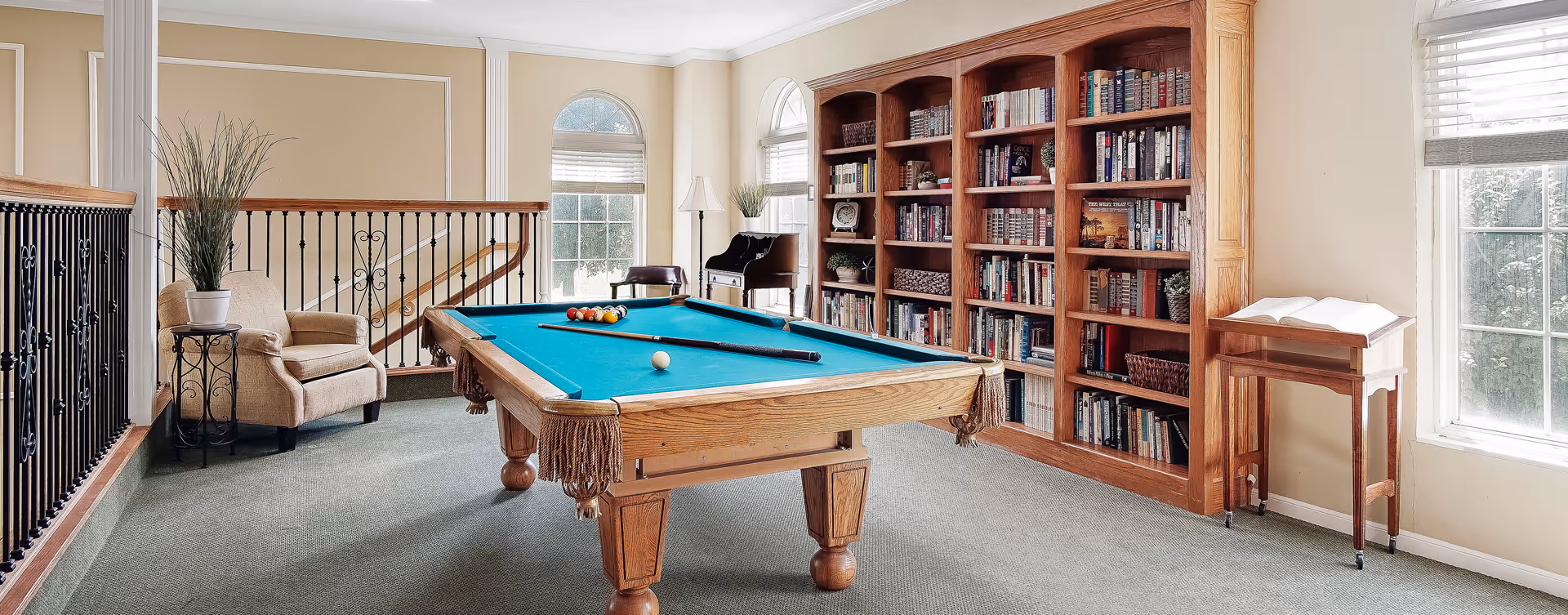 A bright and cozy room featuring a pool table with a blue felt surface and pool balls arranged for a game. The room has large windows with white blinds allowing natural light to fill the space. There is a beige armchair with a potted plant on a small decorative table next to it. A large wooden bookshelf filled with books and decorative items lines one wall. The room has light-colored walls and carpeted flooring, creating a comfortable and inviting atmosphere.