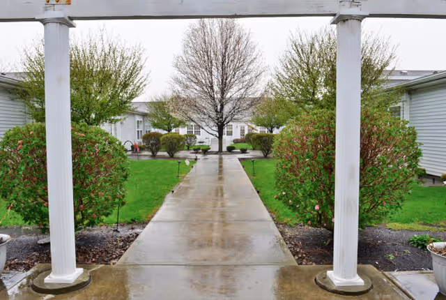 View of a wet concrete walkway lined with green bushes and small trees, leading to single-story buildings under an overcast sky. The walkway is framed by white columns and a pergola structure at the front.