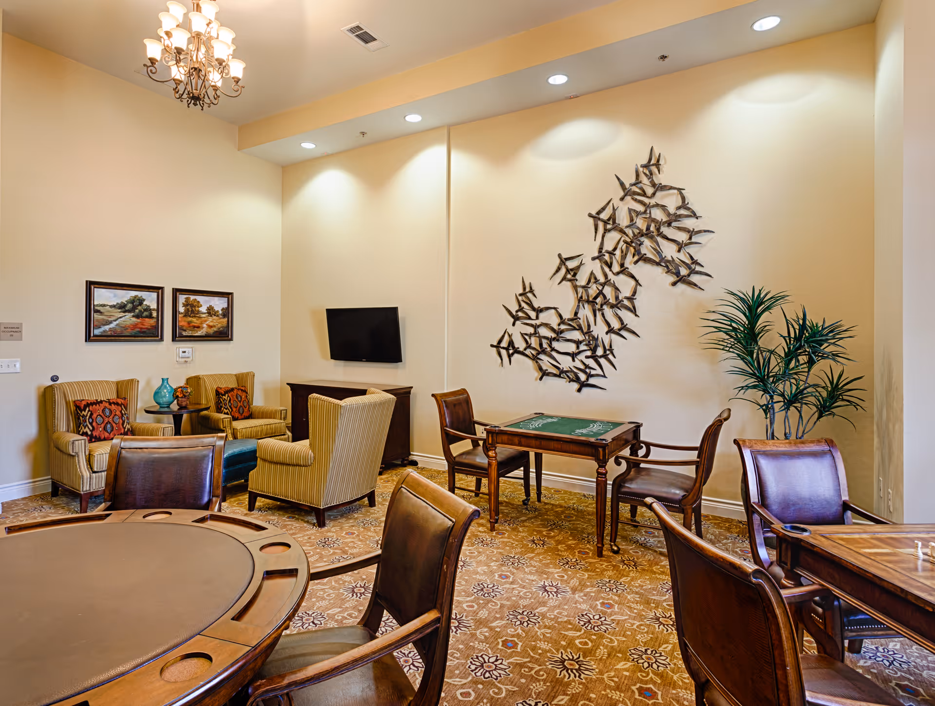 A cozy common area in an assisted living facility featuring a round poker table with chairs, a smaller table with two chairs, and a seating area with four armchairs and a small round table. The walls are decorated with two landscape paintings and a metal wall art piece resembling a flock of birds. A flat-screen TV is mounted on the wall, and a potted plant adds greenery to the space. The room is warmly lit with a chandelier and recessed ceiling lights.