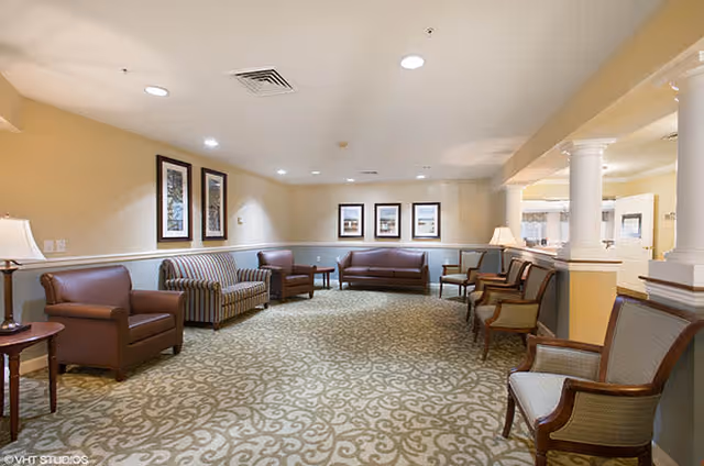 A spacious, well-lit common area with patterned carpet, beige walls, and recessed lighting. The room features multiple seating options including leather armchairs, striped and solid sofas, wooden chairs with cushions, side tables with lamps, and framed artwork on the walls. White columns partially separate the seating area from an adjacent space.