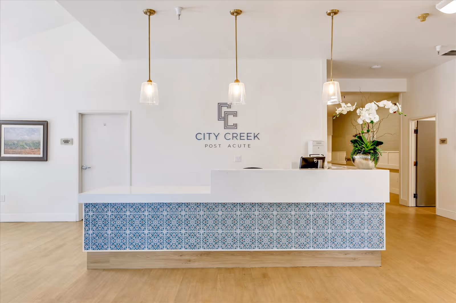 Reception area of City Creek Post Acute facility featuring a white front desk with blue patterned tiles on the front, three pendant lights hanging from the ceiling, a potted orchid plant on the desk, and the facility's logo on the wall behind the desk.
